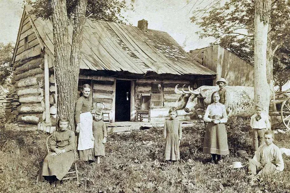 Familia de colonos frente a una cabaña de troncos con techo de tejas de madera, un porche de tablones y un buey, fines del siglo XIX