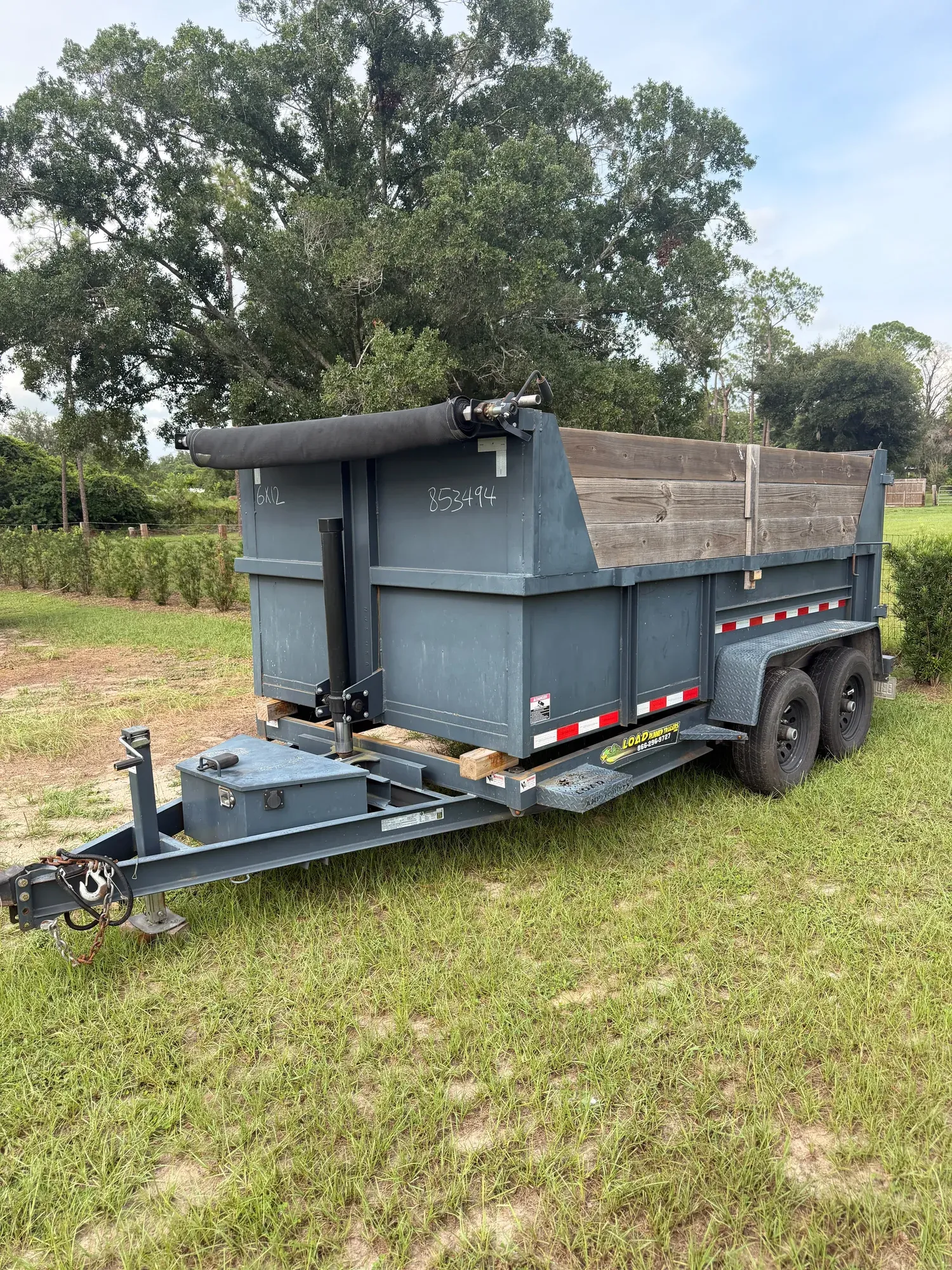 Grey dump trailer parked on grass, seen from the side, empty with its wooden side boards up and the tarp rolled back