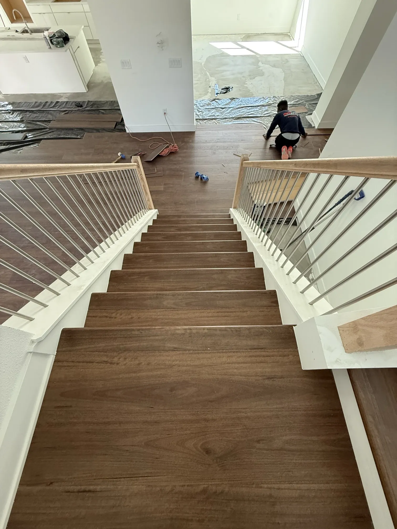 Installer kneeling to lay plank flooring at the top of a newly finished staircase, seen from the landing above