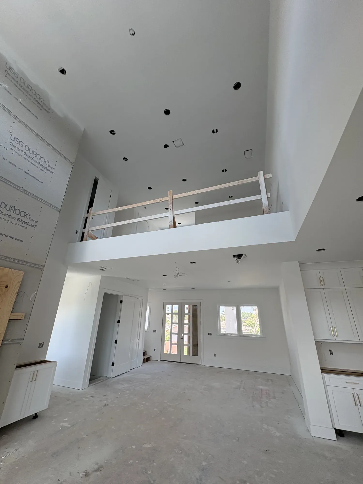 Double-height great room under construction, with a bare concrete slab, a temporary timber railing on the loft and cement board sheets on the far wall