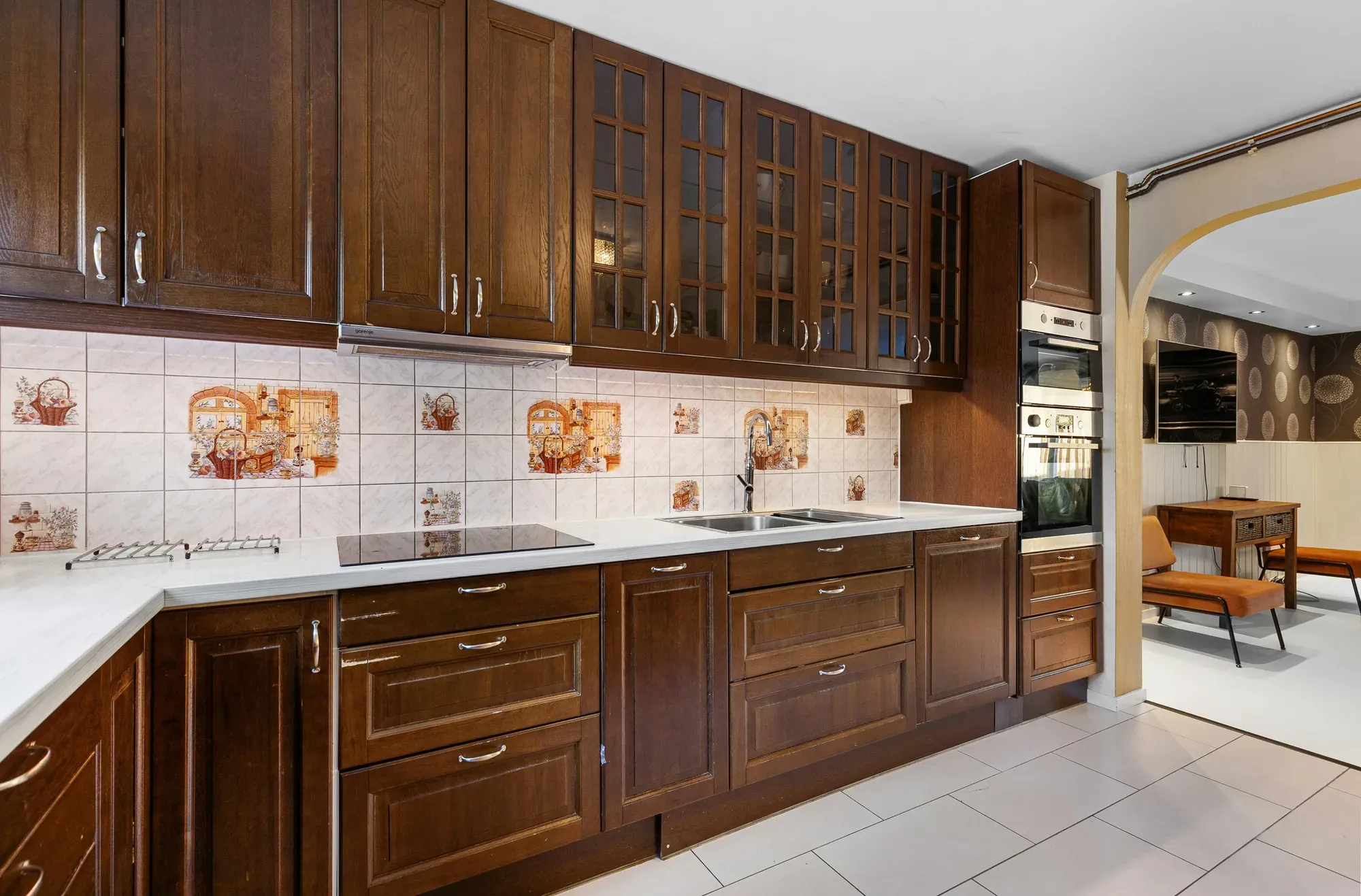 A dark wood kitchen with glass-front upper cabinets, a light countertop, a built-in cooktop and a patterned tile backsplash