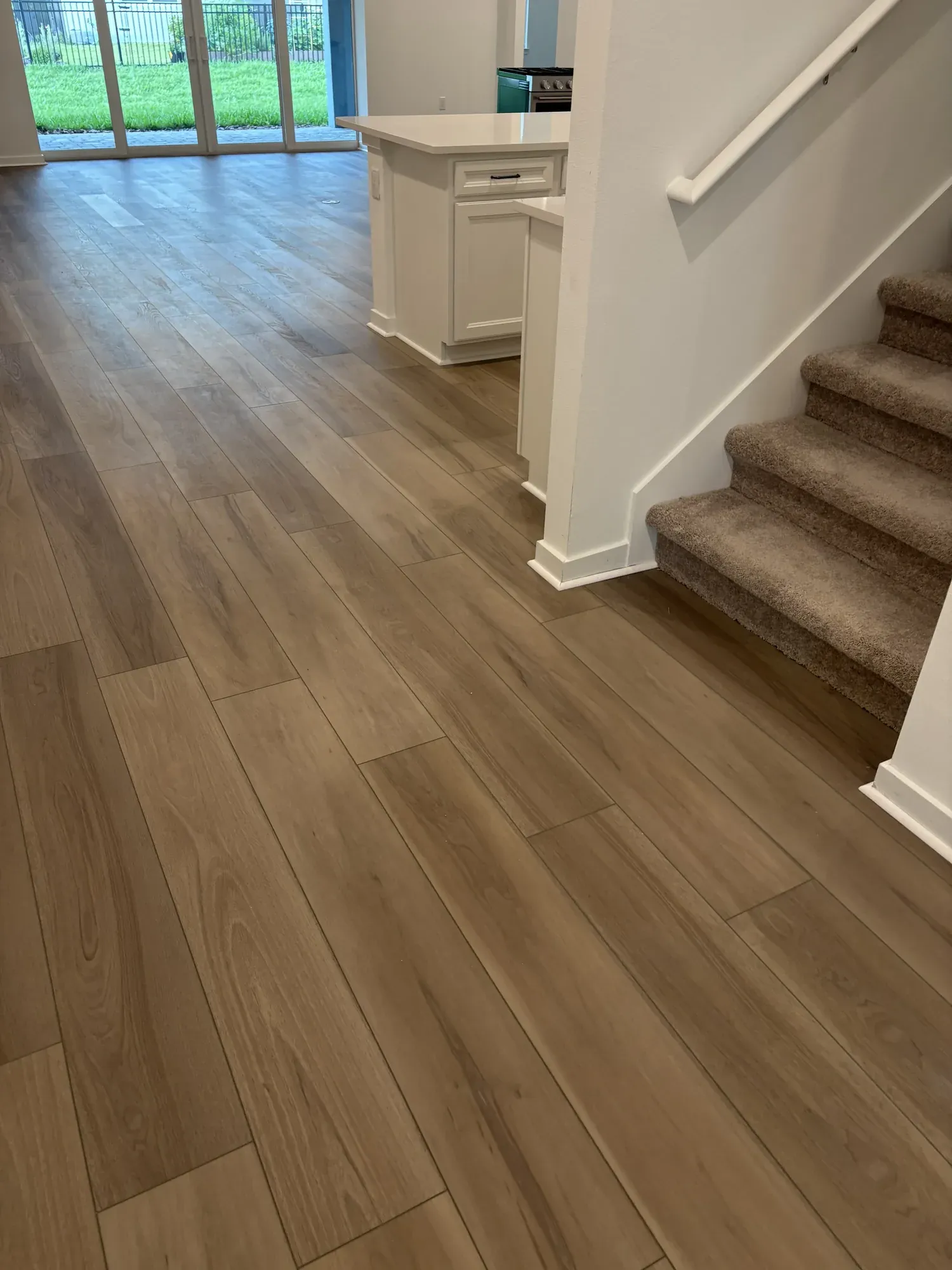 Empty finished home with wide wood-look plank flooring running from the entry past a white kitchen island, with a carpeted staircase to the right and sliding doors onto a lawn