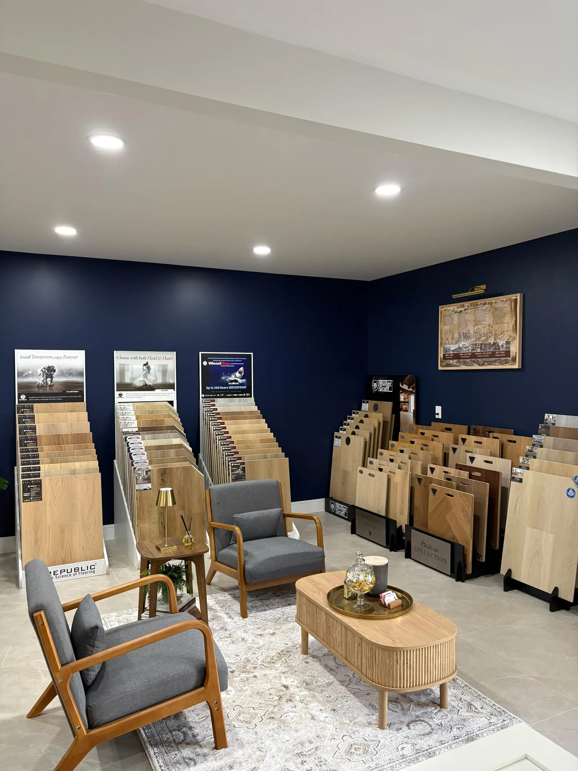 The showroom seating area seen from the entrance: rows of flooring sample boards on stands along the navy wall, two gray armchairs and a round wooden table on a rug